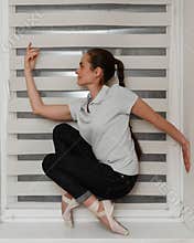 A female ballerina in casual clothes in jeans and a t-shirt in Pointe shoes at home on the windowsill posing for the camera. The