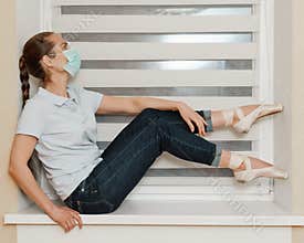 A female ballerina in a mask in casual clothes in jeans and a t-shirt in Pointe shoes at home on the windowsill posing for the
