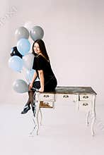 Happy emotional glamour smiling young woman in black dress and shoes with birthday holiday ballons sitting on white