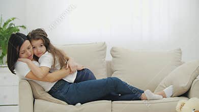 Cute little kid girl and lovely mother are lying on the sofa, smiling, kissing, and hugging each other.