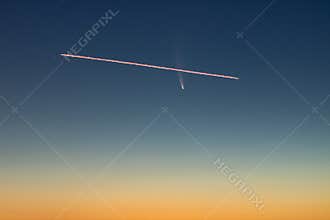 A plane flying in front of C/2020 F3, or Comet Neowise, in the early morning sky with a gradient from orange sun glow to blue nigh