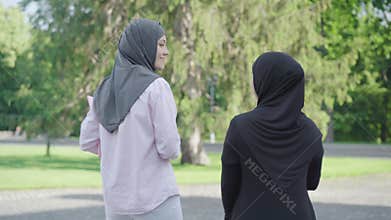 Back view of positive immigrants with books walking outdoors on sunny day and talking. Two happy Muslim women in hijabs