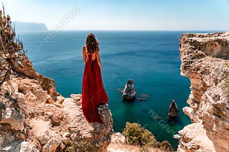 From behind, a woman is seen in a red flying dress fluttering in the wind against the background of a sea with rocks