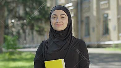 Young proud Muslim woman in traditional black hijab posing on sunny day at university yard. Portrait of confident female