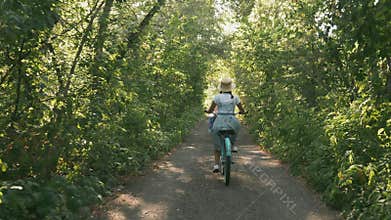 Girl cyclist is riding retro bike in green park in sunlight. Woman in hat rides old bicycle on forest road at sunrise. Happy femal