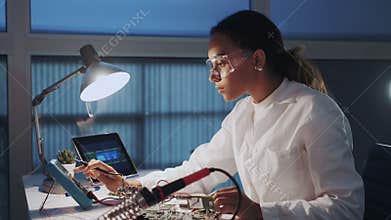 Middle close up of female engineer of electronics in white coat and protective glasses checking motherboard with
