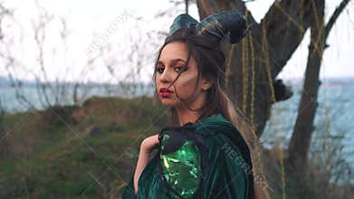 Bloody witch, girl with big black horns on head in green emerald mantle, lady with dark hair and brown eyes looks at