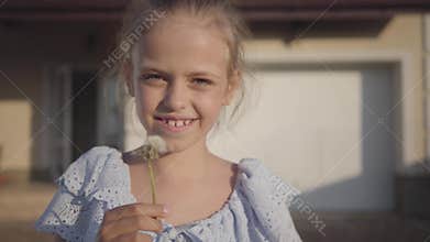 Portrait of a pretty cute little girl blowing a dandelion and looking at the camera smiling. The child spending time