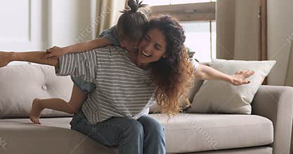 Happy mum playing piggybacking cute kid daughter sit on couch