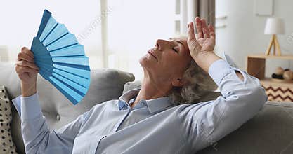 Overheated exhausted old woman feel hot wave fan on sofa