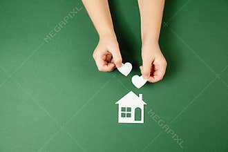 Family home and real estate concept. Close up of child hands holding white paper house and heart on green background.