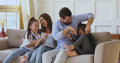 Smiling mum and dad tickling little children playing on sofa