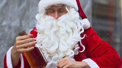 Bad drunk Santa Claus sits near a city office building and uncovers a bottle of alcohol. Santa Claus reads the alcohol