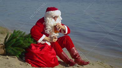 Santa Claus sits on the sandy shore of the lake sniffing the smell of alcohol and bottles. Drunk Santa Claus