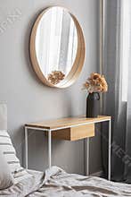 Elegant round mirror in wooden frame above fancy console table with flowers in vase in trendy bedroom interior with beige vase