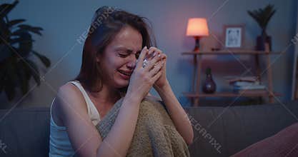 Close up of lonely millennial female in tears sitting on couch in fetal position covered with banket . Young woman
