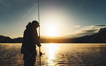 Silhouette fisherman wirh fishing rod at sunrise sunlight, outline man enjoy hobby sport on evening lake, person catch fish