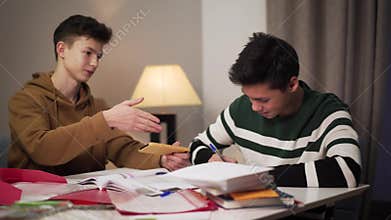 Teenage Caucasian boy distracting his Asian groupmate from studies. Two male college students sitting at the table with