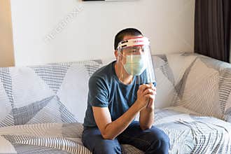 Stressed young Asian man with mask and face shield praying while watching tv at home under quarantine