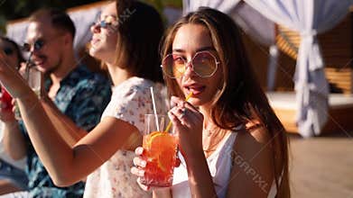 Pretty woman in stylish sunglasses toasting with friends, drinking cocktail of plastic straw at pool party. Group of