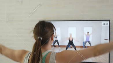 The girl does exercises while watching a program on TV at home. A young woman in sportswear, athletic build, plays