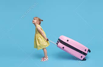 Cute baby girl in a summer dress carries a suitcase. waiting for travel after quarantine. Blue studio background.