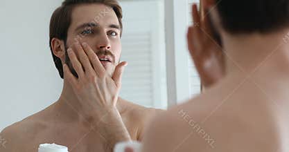 Charming young happy man applying moisturizing facial creme.