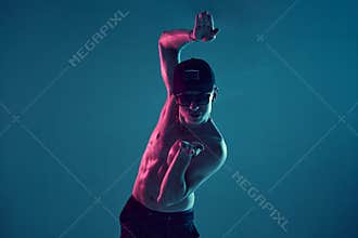 Young guy in cap and sunglasses dancing break dance without t-shirt in studio in blue neon light. Break dance lessons