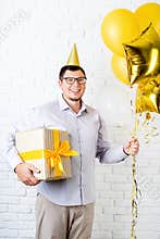 Funny young man wearing birthday hat holding balloons and a big gift box celebrating birthday over white brick background