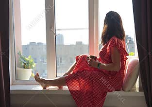 A woman sits on a window seat in the summer sun and looks out at the street
