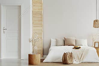 Flower on glass vase on round wooden table in beige natural bedroom, copy space on empty wall