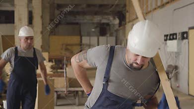 Portrait of adult Caucasian man in white protective helmet walking at the construction site with a wooden plank. Builder