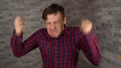 A young handsome man in a plaid shirt freaks out and waves his hands on a gray brick background.