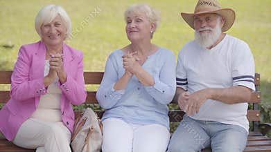 Three caucasian middle-aged people sitting at the bench and applauding. Elderly company having fun in the park after