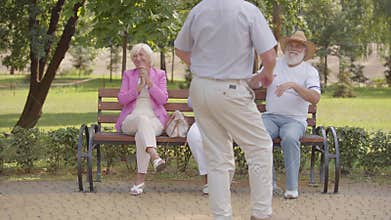 Three caucasian seniors sitting at the bench and applauding their cheerful male friend dancing. Elderly company having
