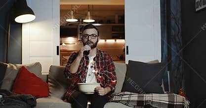 Young handsome geeky Caucasian student man watching movie alone at home with party lights eating snacks slow motion.