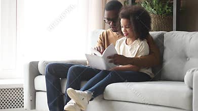 African father reading book to daughter sitting together on couch
