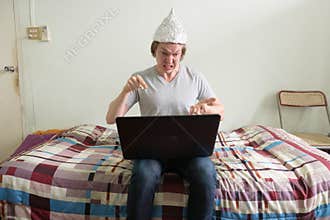 Stressed young man with tin foil hat using laptop and looking angry in the bedroom