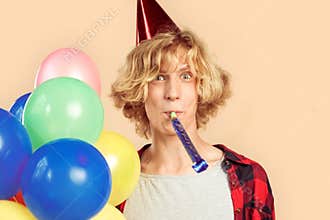 Freestyle. Teen blond boy in birthday cap standing isolated on color with balloons blowing party horn smiling