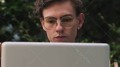 Closeup of serious young man working on laptop computer in cafe outdoors. Man being deep in thought