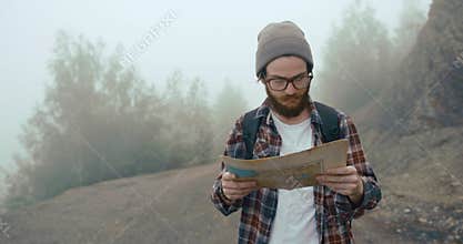 Traveling in the mountains. Young man with stylish beard learns an old map walking on the mountain covered with thick