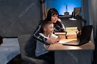mom helping son with homework while sitting at desk at home using laptop and books at night. online schooling