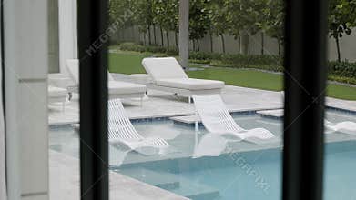 View from window on lounge area near inground swimming pool with six white deck chairs. Interior design of splendid