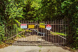 Old metal gate on shady road with chains and locks and signs saying no entry security cameras in use and no trespassing
