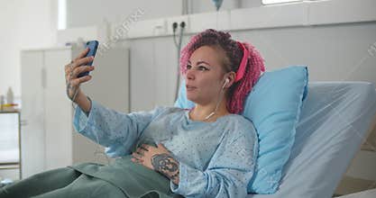 Portrait of sick woman speaking by video call in hospital and waving to camera lying in bed