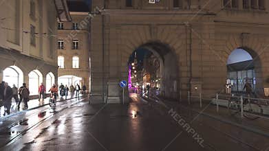 The center of Bern at night with buses, trams, statues and cathedrals with locals and tourists walking by