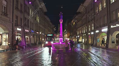 The center of Bern at night with buses, trams, statues and cathedrals with locals and tourists walking by