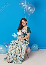 Pregnant cheerful brunette young woman sitting on the chair with balloons, on blue background.