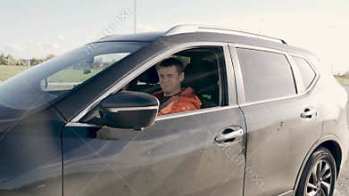 The driver is driving a car, waving his hand in greeting