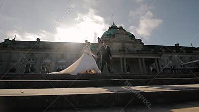Bride and groom holding hands on stone steps in front of historic building, elegant wedding scene in sunlight
